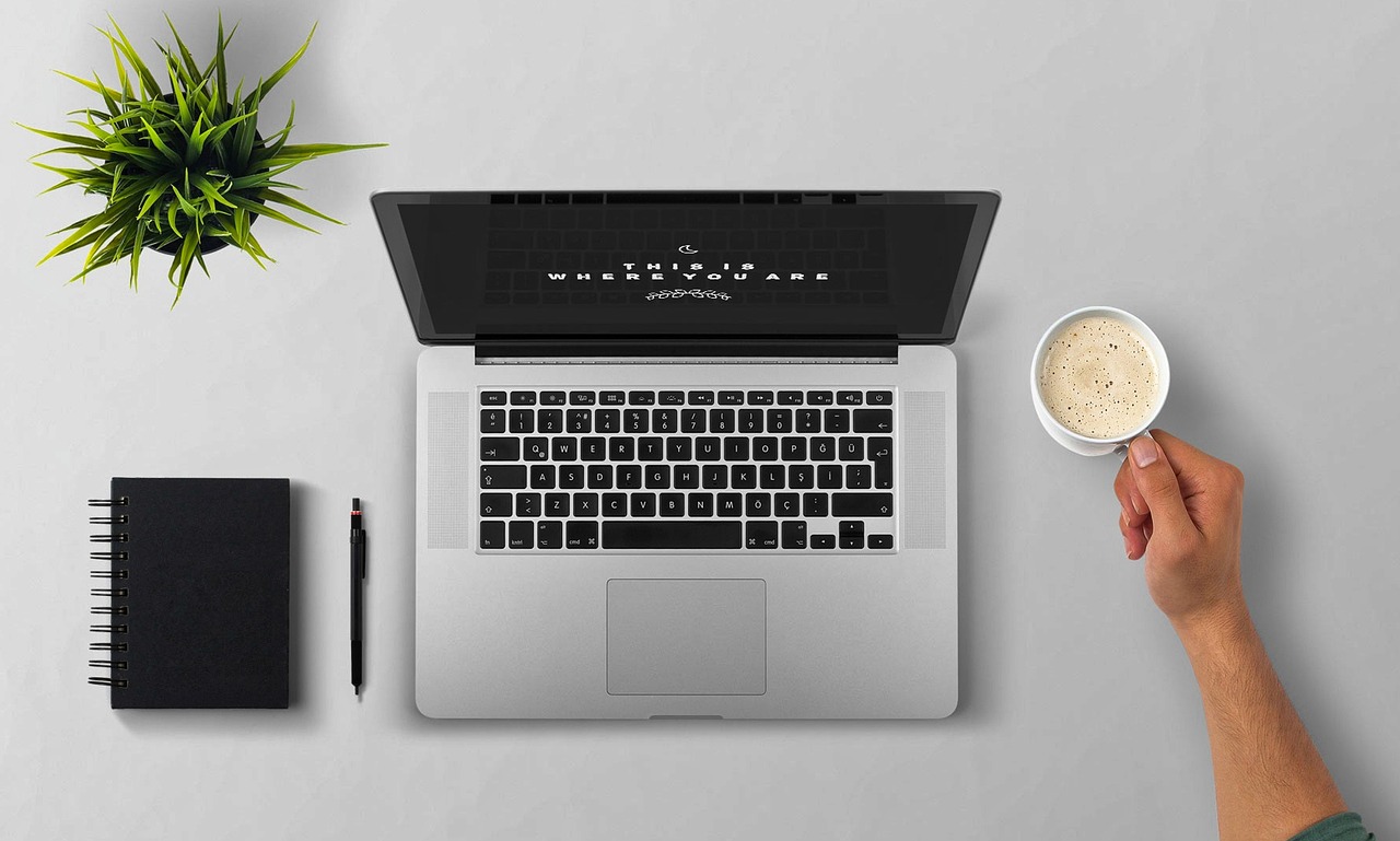 Imagen de una notebook junto a una mano sosteniendo una taza de café sobre un escritorio. Esta imagen evoca un ambiente laboral productivo y acogedor, ideal para realizar tareas de consultoría con comodidad.